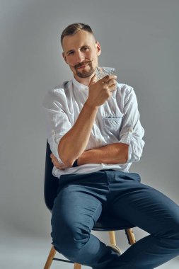 Klasik beyaz tişörtlü ve mavi pantolonlu şık bıyıklı bir adam sandalyeye oturmuş, elinde bir bardak viski tutuyor. Gri arkaplan, yakın çekim.