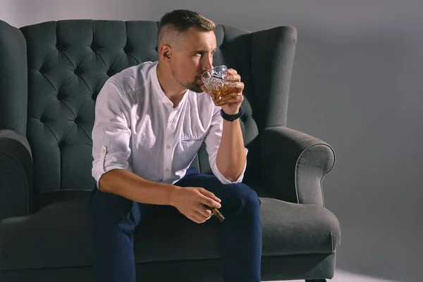 Muž se stylovým knírem, oblečený v bílé košili, modré kalhoty sedí na tmavé pohovce, drží sklenici whisky, doutník. Šedé pozadí, detail. — Stock fotografie