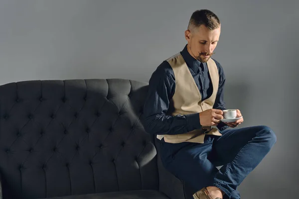 Hombre con mostache elegante, vestido con camisa negra y pantalones, beige vest se sienta en un sofá oscuro, tomando una taza de café. Fondos grises y cierres. — Foto de Stock