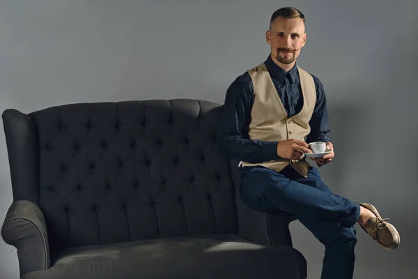 Hombre con mostache elegante, vestido con camisa negra y pantalones, beige vest se sienta en un sofá oscuro, tomando una taza de café. Fondos grises y cierres. — Foto de Stock