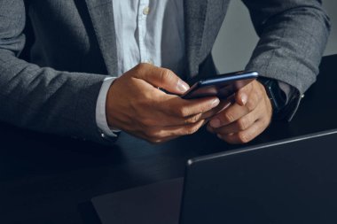 Klasik takım elbiseli, beyaz gömlekli, ofiste masada oturan, akıllı telefonunu kullanan, gri arka planlı iş adamının elleri. Yakın plan..