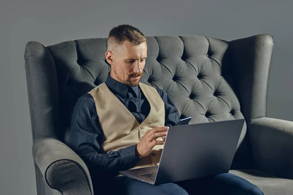 Mann mit stylischem Schnurrbart, schwarzem Hemd und beiger Weste sitzt auf dunklem Sofa und bedient Laptop und Smartphone. Grauer Hintergrund, Nahaufnahme. — Stockfoto