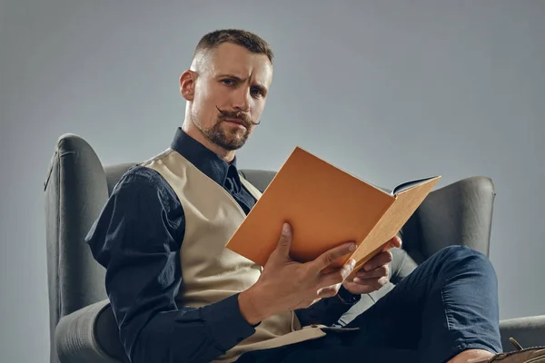 Άνδρας με κομψό μουστάκι, ντυμένος με μαύρο πουκάμισο και παντελόνι, μπεζ γιλέκο κάθεται σε σκούρο καναπέ, διαβάζοντας ένα περιοδικό. Γκρι φόντο, κοντινό πλάνο. — Φωτογραφία Αρχείου