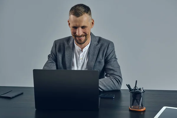 El empresario portador de mostache con estilo, vestido con un traje clásico, está trabajando en el ordenador portátil mientras se sienta a la mesa en la oficina, fondo gris.. — Foto de Stock