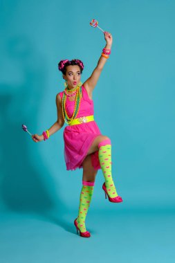 Çok renkli saç stili ve parlak makyajlı güzel bir kız. Stüdyoda mavi arka planda poz veriyor. Elinde lolipop tutuyor..