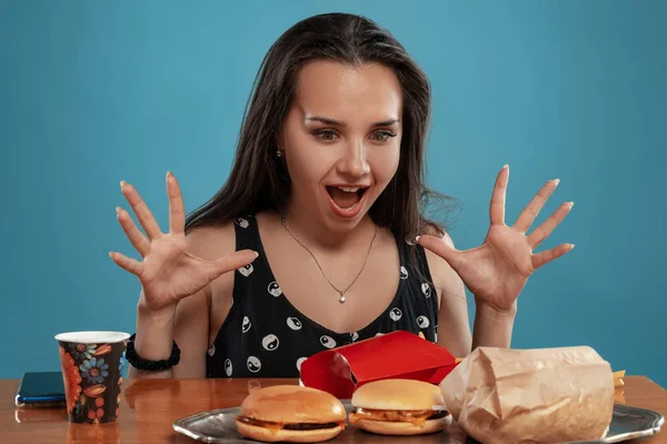 Portret kobiety w czarnej sukience, siedzącej przy stole z hamburgerami, frytkami i piciem. Niebieskie tło. Fast food. — Zdjęcie stockowe