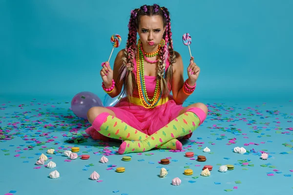 Chica encantadora con un peinado de trenzas multicolores y maquillaje brillante, posando en el estudio con piruleta, globos aéreos y confeti sobre un fondo azul . —  Fotos de Stock