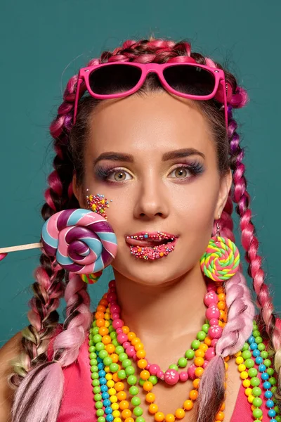 Menina encantadora com um penteado de tranças multi-coloridas e maquiagem brilhante, posando em estúdio contra um fundo azul, segurando um pirulito em sua mão . — Fotografia de Stock