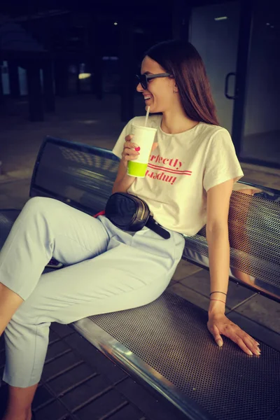 Retrato de menina em óculos escuros posando sentado no banco no shopping center. Vestido em t-shirt branca, calças azuis, bolsa de cintura preta. — Fotografia de Stock