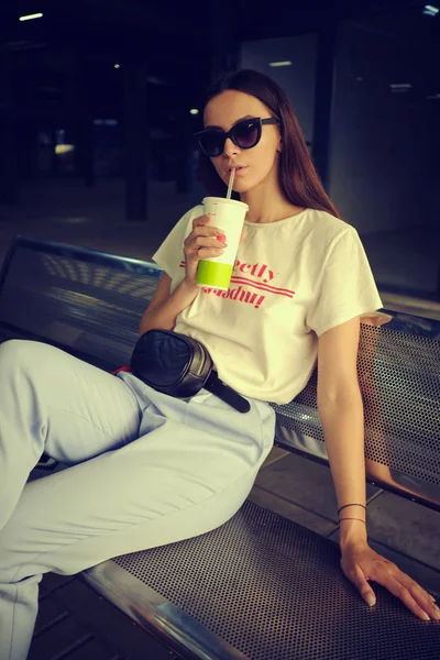 Portrait of girl in dark sunglasses posing sitting on bench in shopping mall. Dressed in white t-shirt, blue trousers, black waist bag. – stockfoto