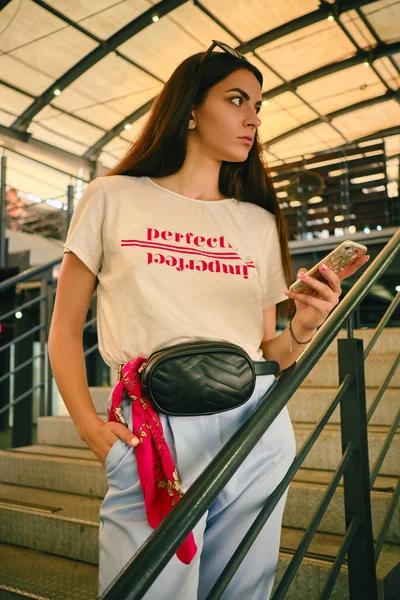 Портрет девушки, позирующей возле торгового центра, держащей в руках смартфон. Был одет в белую футболку, синие брюки, черную сумку.. — стоковое фото