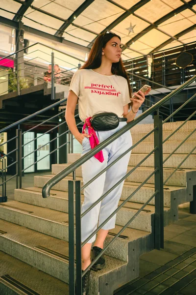 Porträt eines Mädchens, das in der Nähe eines Einkaufszentrums posiert und sein Smartphone in der Hand hält. Bekleidet mit weißem T-Shirt, blauer Hose, schwarzer Taille und Turnschuhen. — Stockfoto