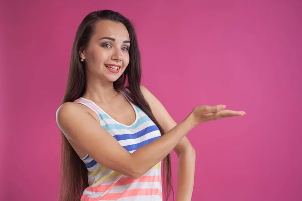 Mulher morena com cabelos longos, vestida com camisa listrada colorida, posando contra fundo de estúdio rosa. Emoções sinceras. Close-up. — Fotografia de Stock