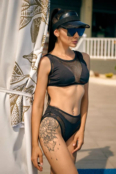 Portrait of a woman having rest and posing near a swimming pool. Dressed in a black swimsuit, sun visor and sunglasses. — Stock Photo, Image