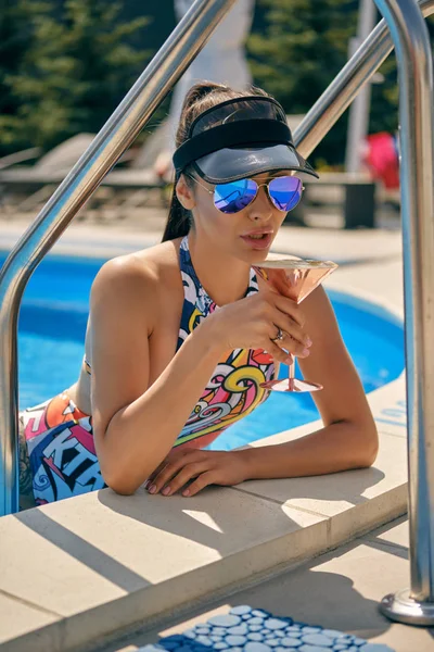 Portrait of a girl having rest and posing near a swimming pool. Dressed in a colorful swimsuit, sun visor and sunglasses. — Stock Photo, Image