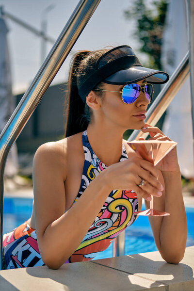 Portrait of a girl having rest and posing near a swimming pool. Dressed in a colorful swimsuit, sun visor and sunglasses.