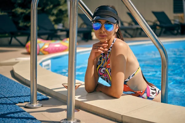Portrait of a girl having rest and posing near a swimming pool. Dressed in a colorful swimsuit, sun visor and sunglasses. — Stock Photo, Image