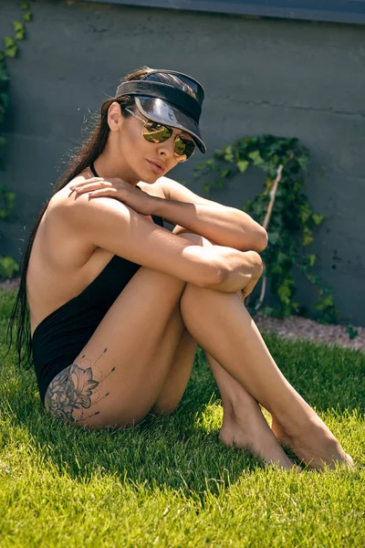 Woman posing sitting on a green grass of a recreation center or hotel with a swimming pool. Dressed in a black swimsuit, sun visor and sunglasses. — Stock Photo, Image