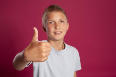 Beyaz tişörtlü yakışıklı sarışın bir gencin yakın plan portresi. Gülümsüyor ve pembe bir stüdyoda poz verirken başparmağını kaldırıyor. Samimi duygular, mutlu çocukluk kavramı.