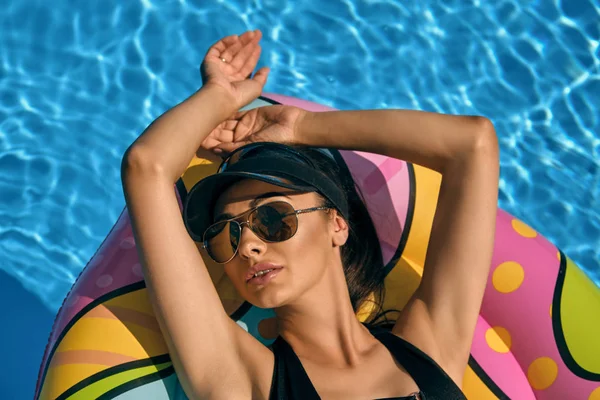 Портрет жінки, яка відпочиває і позує в басейні на надувному матраці. Одягнений у чорний купальник, шапка для сонця та сонцезахисні окуляри . — стокове фото