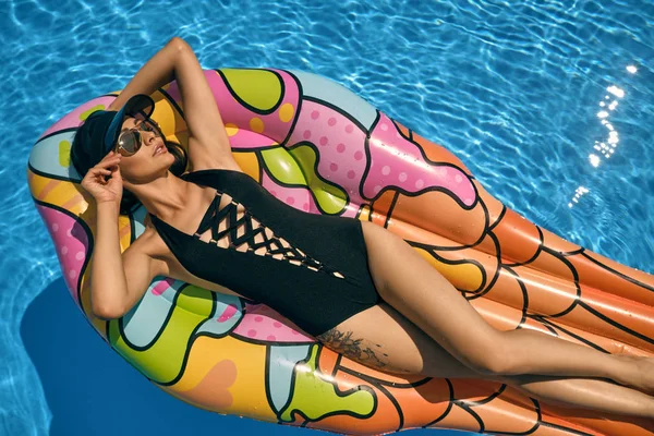 Portret van een vrouw die rust heeft en poseert in een zwembad op een opblaasbaar matras. Gekleed in een zwart badpak, zonneklep en zonnebril. — Stockfoto