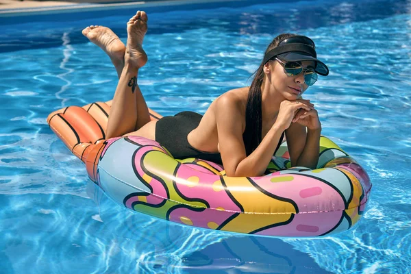 Портрет жінки, яка відпочиває і позує в басейні на надувному матраці. Одягнений у чорний купальник, шапка для сонця та сонцезахисні окуляри . — стокове фото