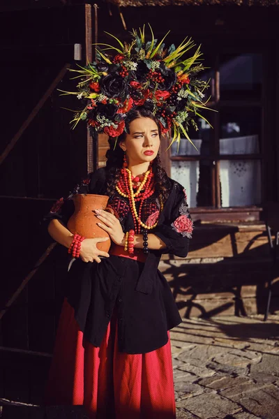 Bruna ragazza in un nero e rosso ucraino ricamato autentico costume nazionale e una corona di fiori posa in piedi al cancello . — Foto Stock