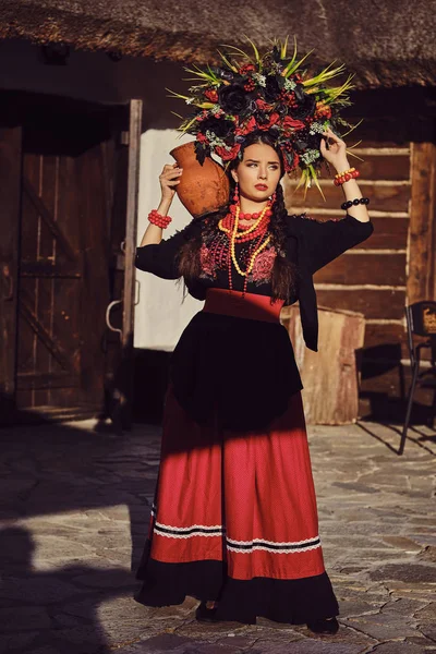 Bruneta fata într-un negru și roșu ucrainian brodat autentic costum național și o coroană de flori se prezintă în picioare la poartă . — Fotografie, imagine de stoc