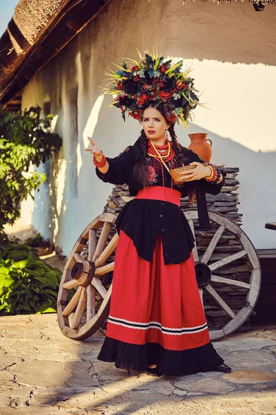 Brunett flicka i svart och rött broderade ukrainska autentiska nationella kostym och en krans av blommor poserar stående mot en vit koja. — Stockfoto