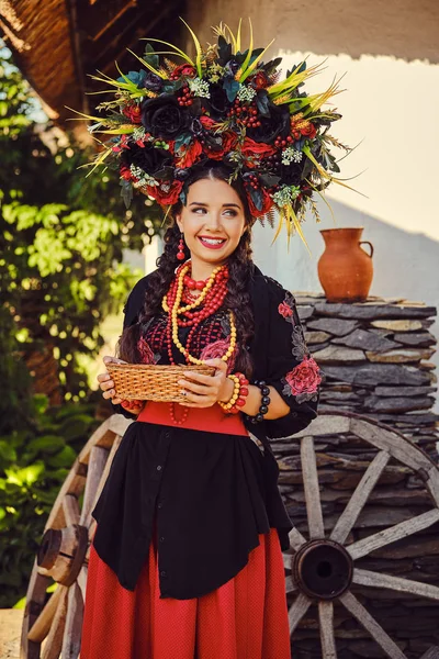 Brunetka dziewczyna w czerni i czerwieni haftowane ukraiński autentyczny strój narodowy i Wieniec kwiatów pozuje stoi przed białą chatę. — Zdjęcie stockowe