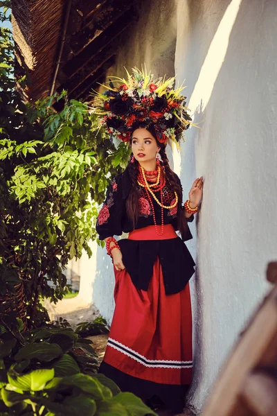 黒と赤の刺繍ウクライナの本格的な民族衣装と花輪のブルネットの女の子は、白い小屋に立ってポーズをとっています. — ストック写真