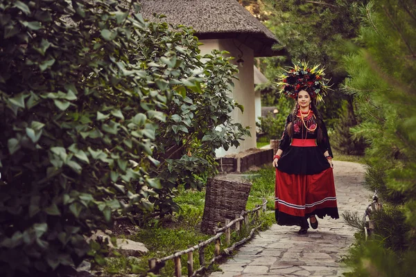 Bruna ragazza in nero e rosso ricamato ucraino autentico costume nazionale e corona di fiori sta camminando da solo e in posa in un giardino verde . — Foto Stock