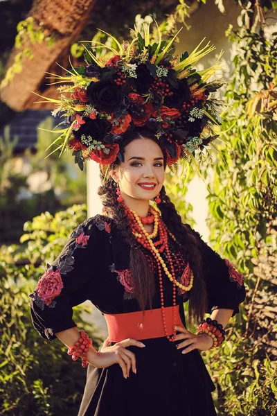 Bruna ragazza in nero e rosso ricamato ucraino autentico costume nazionale e una corona di fiori posa in piedi contro una capanna bianca . — Foto Stock