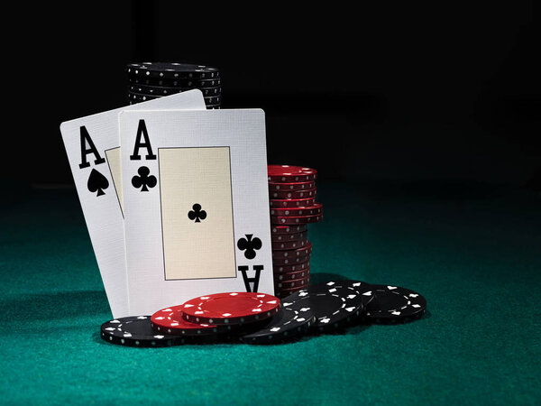 Two aces standing leaning on chips piles, some of them laying nearby on green cover of playing table. Black background. Close-up.
