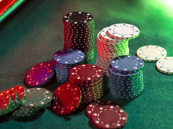 Vista superior de cerca de las pilas de chips de colores, algunos de ellos colocados cerca en la cubierta verde de la mesa de juego, bajo la luz de neón verde, rojo. Fondo negro . — Foto de Stock
