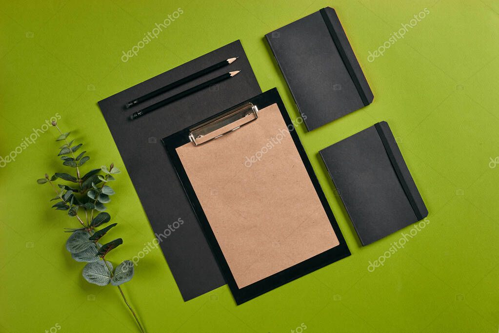 Sheet of black paper, clipboard with brown paper, pencils, folders and a twig on green background with copy space. Education, workplace concept. Top view in focus with different office equipment, supplies, stationery. Close-up shot.