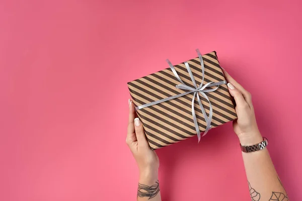 Manos de una mujer con tatuajes y ver la celebración de la caja de regalo marrón rayado atado con cinta de plata en un fondo rosa. Cierre, espacio de copia, vista superior. —  Fotos de Stock