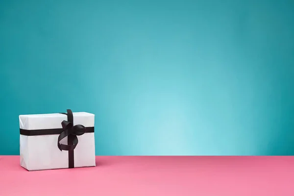 Confezione regalo in carta bianca con nastro nero e fiocco su una superficie rosa e sfondo blu. Primo piano, copiare lo spazio. — Foto Stock