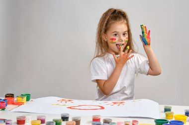 Beyaz tişörtlü küçük bir kız masasında hangi adamla oturuyordu ve üzerine resim çiziyordu, ellerindeki boyaları yalıyordu. Beyazda izole edilmiş. Orta boy yakın çekim.