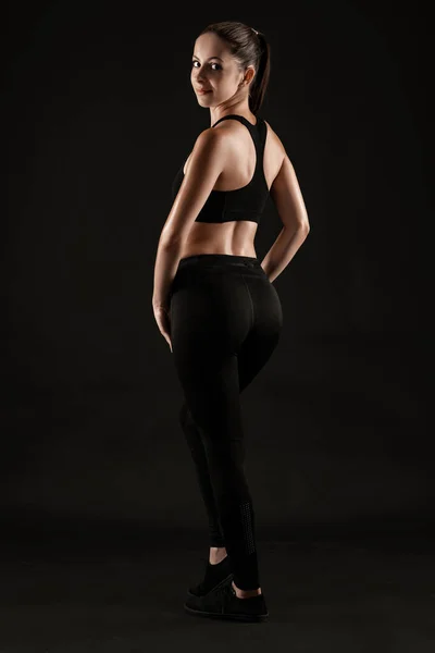 Mulher morena em leggings pretos, top e tênis está posando contra um fundo preto. Fitness, ginásio, conceito de estilo de vida saudável. Comprimento total. — Fotografia de Stock
