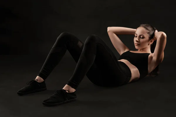 Mulher morena em leggings pretos, top e tênis está posando contra um fundo preto. Fitness, ginásio, conceito de estilo de vida saudável. Comprimento total. — Fotografia de Stock