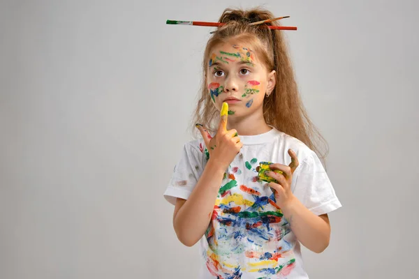 Little girl in white t-shirt, with brushes in her hair is posing standing isolated on white, gesticulating with painted hands and face. Close-up. — Stock Photo, Image