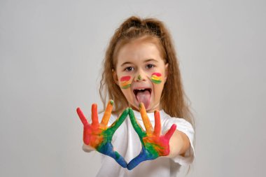 Beyaz tişörtlü küçük bir kız beyaz üzerinde tek başına poz veriyor ve farklı renklerde resimleriyle el kol hareketleri yapıyor. Sanat stüdyosu. Yakın plan..