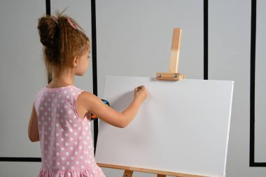 Pembe elbiseli küçük kız ressam resim sehpasının arkasında duruyor ve beyaz duvarlı bir sanat stüdyosunda fırçayla resim yapıyor. Orta boy yakın çekim..