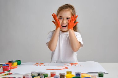 Beyaz tişörtlü küçük bir kız masasında hangi adam ve rengarenk boyalarla oturuyor, yanaklarını boyuyor. Beyazda izole edilmiş. Orta boy yakın çekim.