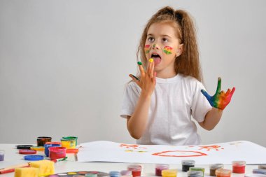 Beyaz tişörtlü küçük bir kız masasında hangi adamla oturuyordu ve üzerine resim çiziyordu, ellerindeki boyaları yalıyordu. Beyazda izole edilmiş. Orta boy yakın çekim.