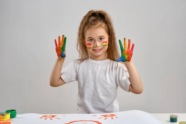 Petite fille en t-shirt blanc debout à table avec whatman et des peintures colorées,, montrant les mains peintes, le visage. Isolé sur blanc. Gros plan moyen. — Photo