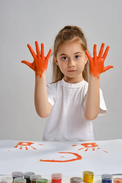 Little girl in white t-shirt sitting at table with whatman and colorful paints, showing her painted hands. Isolated on white. Medium close-up. — Stock Photo, Image