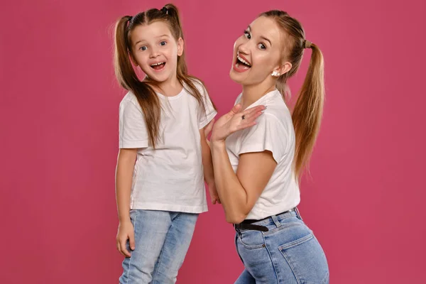 Mãe e filha com um rabo de cavalo engraçado, vestidas com t-shirts brancas e jeans denim azul estão posando contra um fundo estúdio rosa. Close-up shot. — Fotografia de Stock