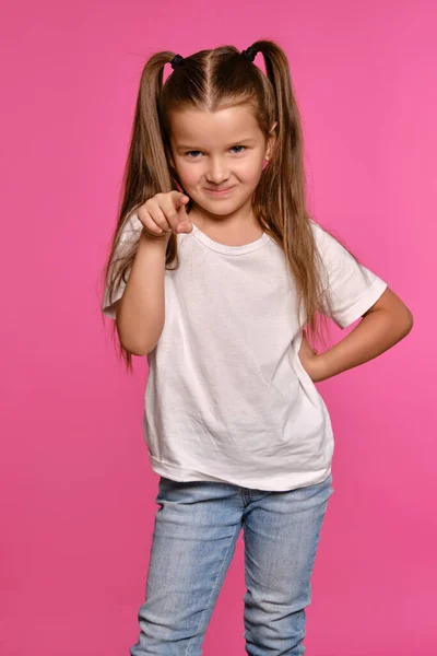 身穿白色T恤和蓝色牛仔裤的马尾辫小女孩是在粉红的工作室背景下摆姿势的。特写镜头。真诚的感情. — 图库照片